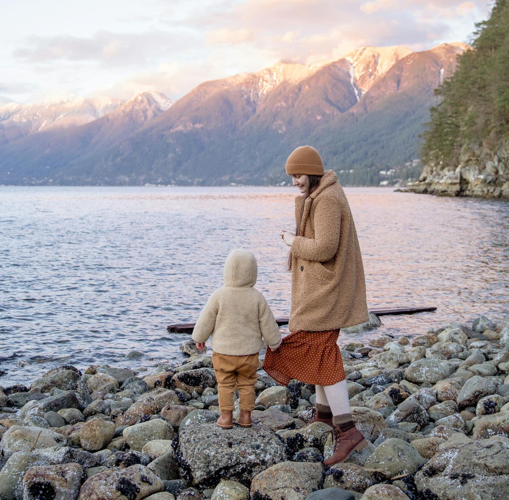 Back view unrecognizable preschool boy in warm outerwear holding skirt of mother in warm clothes and hat while traveling on calm lake in autumn
